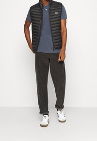 Mann, der eine schwarze gesteppte Weste über einem blauen kurzärmeligen Hemd, dunklen Cordhosen und weißen Sneakers mit schwarzen Streifen trägt.