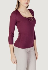 Top fitted bordeaux con maniche tre quarti, con scollatura rifinita in pizzo. Tessuto a costine liscio, abbinato a pantaloni beige.