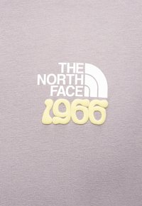 Το λογότυπο The North Face σε λευκό και κίτρινο "1966" σε φόντο από πλεκτό ύφασμα ανοιχτού μοβ χρώματος.