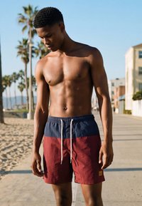 Ung mann iført røde og blå badeshorts står skjorteløs på en solrik strandpromenade med palmer i bakgrunnen.