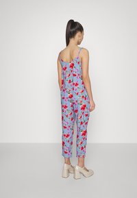 Nainen, jolla on korkea ponihäntä, pukeutuneena siniseen kukalliseen hihattomaan toppiin ja siihen sopiviin cropped-housuihin, sekä béžet korkokengät, seisoo selin.