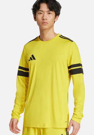 Mladý muž, který nosí žlutý sportovní dres s dlouhým rukávem a černými pruhy na rukávech a ramenou, stojí před jednobarevným pozadím.