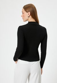 Suéter negro de tela acanalada con mangas largas y cuello alto. Combinado con pantalones blancos, que muestran un diseño ajustado y una superficie texturizada.