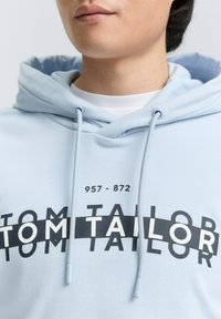 Világoskék kapucnis pulóver, húzózsinórral, fekete grafikai tervezéssel, amely tartalmazza a "TOM TAILOR" márkanevet és számokat. Simább textúra.