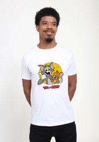 Bílý tričko s krátkým rukávem, které zobrazuje grafiku Toma a Jerryho na žlutém pozadí. Design obsahuje výrazné barvy a hravé kreslené prvky.