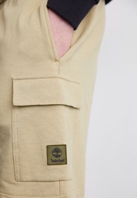 Hand in beige pants pocket above a flap pocket with a green Timberland logo patch on the thigh.