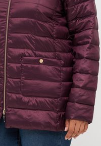 Burgundy puffer jacket with quilted texture, silver zipper, and a snap-button flap pocket, featuring a slightly glossy finish.