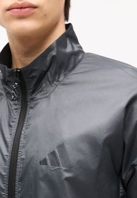 Κοντινό πλάνο ενός ατόμου που φοράει ένα γυαλιστερό σκούρο γκρι μπουφάν με φερμουάρ Adidas, με ψηλό γιακά και λογότυπο στο στήθος.