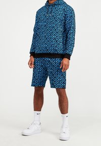 Modrá mikina s kapucí s geometrickými vzory, doplněná o odpovídající kraťasy. Oba kousky mají černé detaily. Nošeno s bílými teniskami a ponožkami.