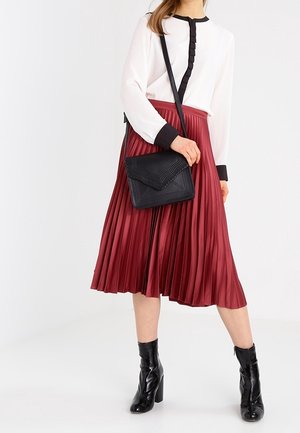 Frau trägt eine weiße Bluse mit schwarzem Rand, einen bordeauxroten plissierten Midi-Rock, schwarze Knöchelstiefel und eine schwarze Umhängetasche vor weißem Hintergrund.