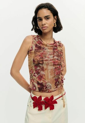 Woman wearing sleeveless floral ruched top and white pants with a red floral belt, standing against a plain light background.
