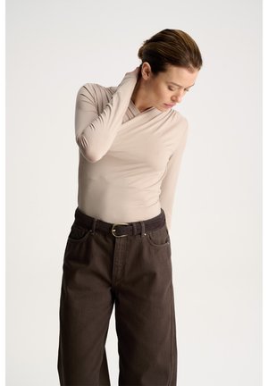Kobieta z włosami upiętymi w kok nosi beżowy długi rękaw i ciemnobrązowe spodnie, przechylając głowę i dotykając szyi na jednolitym tle.