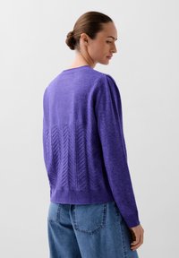 Lilla strikket sweater med et struktureret chevron-mønster på ryggen, ribbet manchetter og kant. Parret med blå denimbukser.