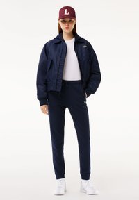Marine bomberjakke, hvit t-skjorte, matchende joggebukser og hvite sneakers. Burgunder caps med 'L'-logo. Fokus på uformell, koordinert sportsbekledning.