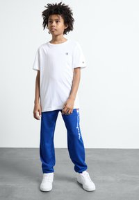 White short-sleeve t-shirt with logo on chest and blue sweatpants featuring white text and logo accents, paired with white sneakers.