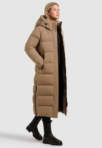 Long, puffy, brown parka with a hood, featuring vertical stitching, side pockets, and a smooth exterior. Paired with black knee-high boots.