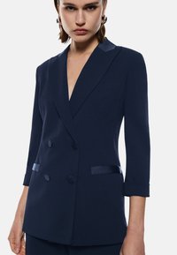 Blazer doppiopetto blu navy con dettagli in raso, spalle strutturate, scollatura a V profonda e maniche a tre quarti. Tessuto morbido.