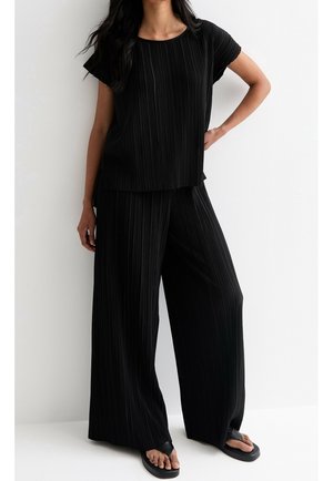 Žena oblečená v černém plisovaném krátkém topu s krátkými rukávy a širokých kalhotách, v kombinaci s černými platformovými sandály, stojící před bílou zdí.