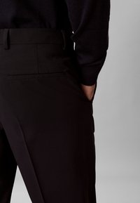 Homme portant un pantalon de costume noir et une chemise noire à manches longues, une main dans la poche latérale, sur fond gris.