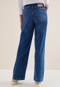 Vaqueros de mezclilla azul de pierna ancha con un acabado ligeramente desgastado, que presentan dos bolsillos traseros, una etiqueta de cuero en la cinturilla y detalles de costura visibles.