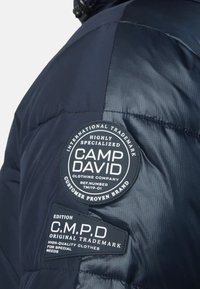 Mörkblå pufferjacka med en slät, glänsande yta. Har ett runt Camp David-logotypmärke och en rektangulär CMPD-etikett.