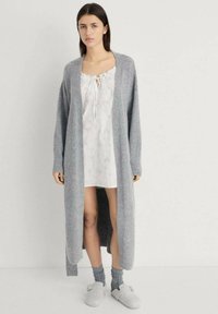 Cardigan lungo grigio realizzato in tessuto morbido, indossato sopra un vestito bianco con scollo a forma di nodo. Calze grigie fino al ginocchio e pantofole leggere completano il look.