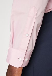 Chemise à manches longues rose clair avec poignets boutonnés, présentant une texture lisse et un ourlet incliné. Les poignets sont dotés de deux boutons blancs pour le maintien.