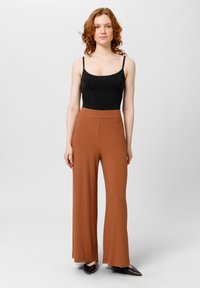 Zwarte bodysuit en ribbelige, roestkleurige wijde broekspijpen. Gladde textuur, hoge taille en een vloeiende pasvorm. Puntige schoenen in donkerbruin.