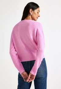 Lys rosa uldtrøje med rund halsudskæring og ribbet kant. Lange ærmer har ribbede manchetter. Båret med blå denim jeans. Bagfra.