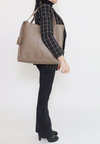 Grote bruine leren tas met een getextureerde afwerking, platte schouderbanden en een klein hangend label als detail. Eenvoudige, rechthoekige vorm.