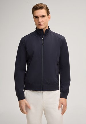 Junger Mann, der eine marineblaue Zip-Jacke und weiße Hose trägt, vor einem schlichten hellen Hintergrund stehend.