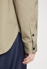 Béžová bavlněná košile s přiléhavým střihem, charakterizovaná zakřiveným lemem, zapínacími manžetami a jemnou texturou. V kombinaci s tmavě modrými proužkovanými kalhotami.