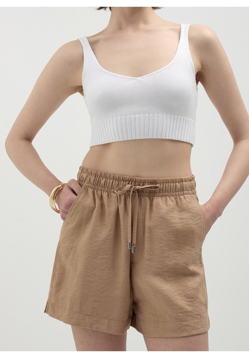 Femme portant un crop top en maille blanche et un short beige à cordon avec des poches, posant les mains dans les poches avec des bracelets en or au poignet.