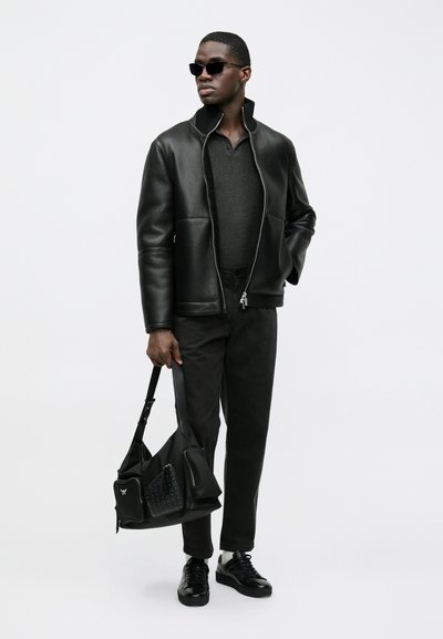 Schwarze Lederjacke mit hohem Kragen, kombiniert mit einem dunkelgrauen Hemd, schwarzen Hosen und Accessoires wie schwarzen Sonnenbrillen und einer Schultertasche.