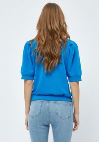 Blå pullovertröja med korta puffärmar, ribbade manchetter och nederkant. Kombinerad med ljusblå tajta jeans, sedd bakifrån.