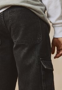 Black denim cargo pants featuring a side pocket, double back pockets, and visible stitching. Material has a textured finish.