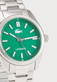 Ceas din oțel inoxidabil argintiu, cu un cadran rotund verde, marcaje albe pentru ore, secundar de culoare violet și logo Lacoste. Are o brățară din metal.