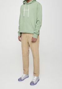 Felpa verde chiaro con tasca frontale, pantaloni beige e sneakers con una parte superiore decorata da un motivo argentato e suole in gomma viola.