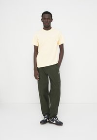 Ανοιχτό κίτρινο T-shirt με μικρό λευκό λογότυπο, συνδυασμένο με σκούρο πράσινο παντελόνι cargo και μαύρα αθλητικά παπούτσια με λευκές λεπτομέρειες.