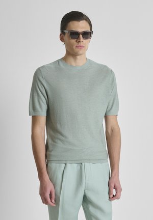 Muškarac obučen u svijetlozeleno pleteno kratkih rukava, odgovarajuće plisirane hlače i tamne pravokutne sunčane naočale, stoji ispred jednobojne pozadine.