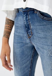 Mano derecha con tatuaje tribal, vistiendo vaqueros azules y una blusa blanca de corte corto y con botones, contra un fondo blanco.