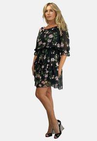 Czarna sukienka w kwiatowy wzór z falbaniastymi warstwami, przezroczystym materiałem, na ramiączkach, z rękawami do łokcia, zestawiona z czarnymi szpilkami.