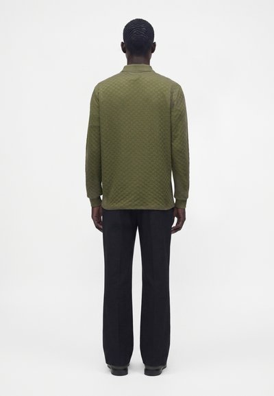 Homme debout de dos, portant une chemise texturée verte à manches longues et un pantalon noir droit sur fond blanc.
