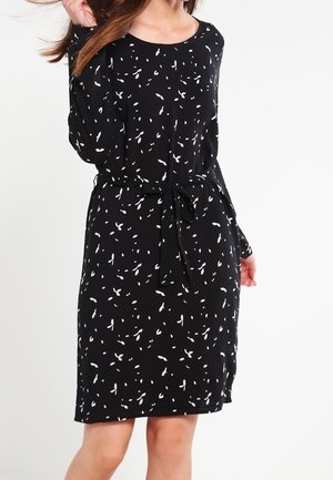 Femme portant une robe noire à manches longues, longueur genou, avec de petits motifs abstraits blancs et une ceinture nouée à la taille.