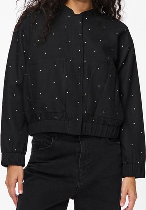 Femme portant une veste courte noire à petits pois blancs, poches avant et poignets côtelés, associée à un jean noir.