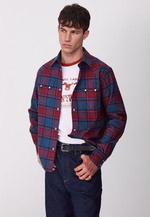 Man draagt een rood-blauw geruit overhemd over een wit T-shirt met afbeelding, donkere spijkerbroek en zwarte riem tegen een effen lichte achtergrond.