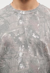 Lähivõte inimesest, kes kannab halli kamuflaažimustriga särki ümar kaelusega.