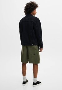 Homme aux cheveux bouclés portant une veste noire, un short vert olive, des chaussettes blanches et des baskets noires, debout de dos sur un fond blanc.