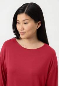 Een vrouw met lang zwart haar in een rode top kijkt lichtjes omlaag en naar de zijkant tegen een plain witte achtergrond.