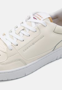 Zapatillas de cuero beige con cordones blancos, detalles de malla texturizada y suela de goma. Presentan un logotipo en relieve en el costado.